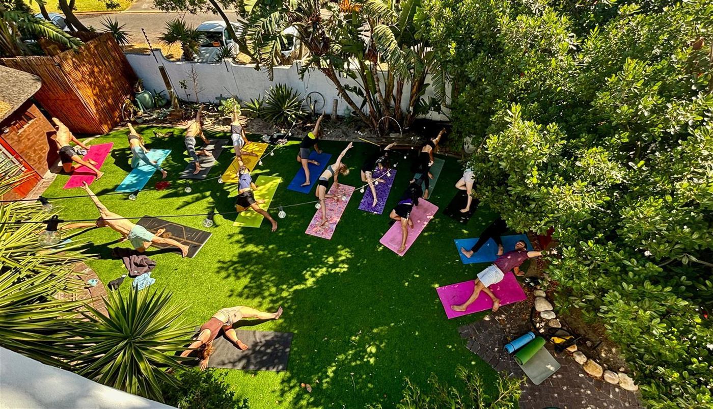Overhead view of a large yoga class on colourful mats across Khaya's green garden lawn in Bloubergstrand, framed by palms and trees